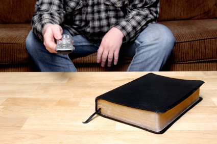 homme assis sur un canapé regardant la télévision, télécommande en main, avec une Bible fermée posée sur la table en face de lui.