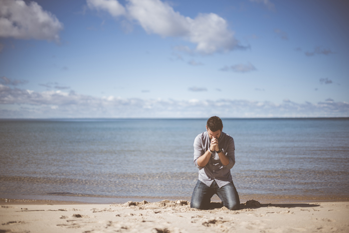 prier dans la nature; homme agenouillé mains jointes en prière, tête inclinée, sur la plage de sable blanc blond, devant la mer calme transparente en fond, sous un beau ciel bleu avec nuages épars