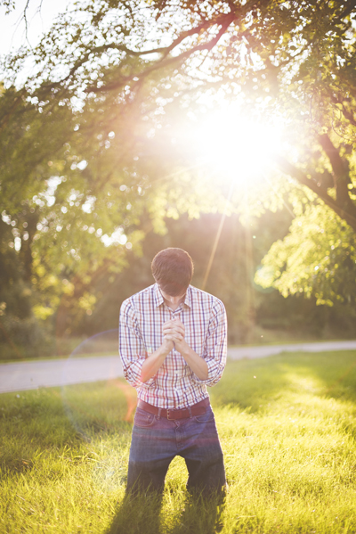 homme en prière, tête inclinée, à genoux, mains jointes, en chemise à carreaux et rayures, jeans bleu, ceinture boucle cuir marron, dans l’herbe ensoleillée du matin, sous les arbres dans la nature, près d’un chemin