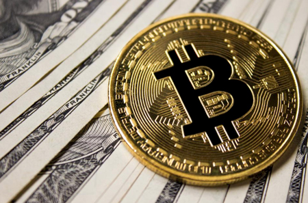 bitcoin; représentation matérielle d’une pièce de crypto-monnaie Bitcoin en or, au-dessus d’une liasse de billets de dollars américains en monnaie fiducière ou monnaie-papier.