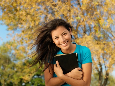 Femme souriante qui tient une Bible contre soi, dans la nature, sur un fond d’automne, arbres feuillus aux couleurs jaunissantes.