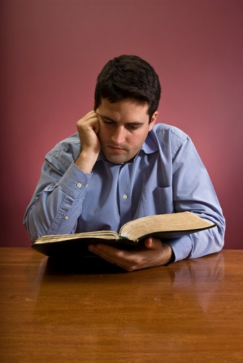 Homme en chemise bleue, lisant la Bible, à une table en bois, sur fond rouge, attitude concentrée