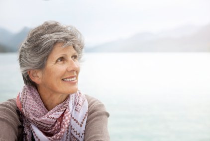 femme âgée souriante, regardant vers le Ciel, cheveux grisonnants, foulard, châle, écharpe, sur un fond d’étendue d’eau, lac, rivière, mer, golfe, baie, rade, et montagnes dans le brouillard