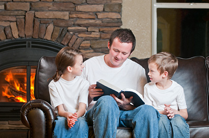 école à la maison; père lisant la Bible à ses deux enfants attentifs, jeans bleu et t-shirt blanc, sur un canapé cuir marron, sur fond de cheminée au bois, foyer allumé, entourée de briques traditionnelles rougeâtres