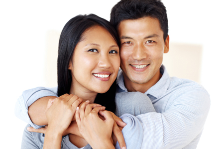 un homme et une femme, couple d’origine asiatique heureux, souriant, cheveux foncés noir, en tenue décontractée mais stylée, le mari enlaçant affectueusement la femme, qui lui répond en posant tendrement ses mains sur son avant bras.