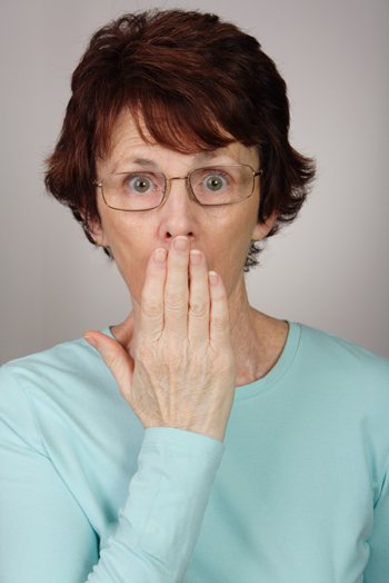 Femme âgée avec des lunettes, cheveux colorés auburn, la main devant la bouche, les yeux écarquillés, étonnée, choquée en entendant parler franchement de la masturbation
