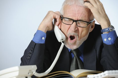 homme d’affaires, patron, en costume noir chemise et montre bleue cravatte foncée, portant des lunettes, cheuveux et barbe grise, fronçant les sourcils, yeux fermés, en colère criant au téléphone filaire blanc, devant un clavier blanc d’ordinateur type apple mac, et un agenda livre ouvert