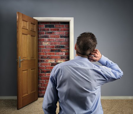 homme vue de dos, se grattant l’oreille, perplexe, regardant une porte en bois ouverte, scellée avec des briques rouges, sur fond de mur peint en couleur grise, ton clair-foncé, encadrure et plinthe blanche, sur moquette beige