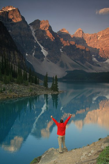 Un homme se réjouissant au bord d'un lac, dont l’eau bleue reflète les montagnes rocheuses alentour, avec des conifères, pins en bordure.