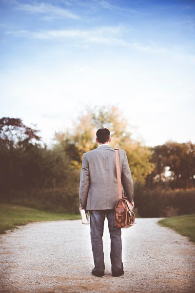 homme de dos marchant sur un chemin dans un parc, une bandoulière sur l’épaule, et une Bible à la main