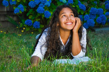fille, jeune femme, étudiant la Bible, allongée dans l’herbe, la tête et les yeux levés vers le Ciel, un large sourire sur le visage, le regard illuminé de joie et de paix, des fleurs bleues et jaunes en arrière-plan, devant un mur boisé