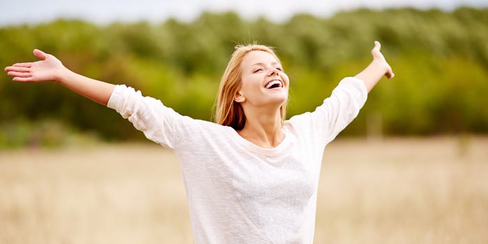 fille, jeune femme heureuse, cheveux blond châtain clair, portant un haut blanc manches longues, souriante à pleines dents, le visage radieux, les bras écartés et mains grandes ouvertes, remerciant et louant le Ciel, dans un champs de céréales à moissonner, une haie végétale arborée en fond