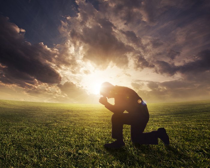 homme agenouillé en prière, tête inclinée sur son poing, dans un champs d'herbe verte, sous le soleil transperçant les nuages