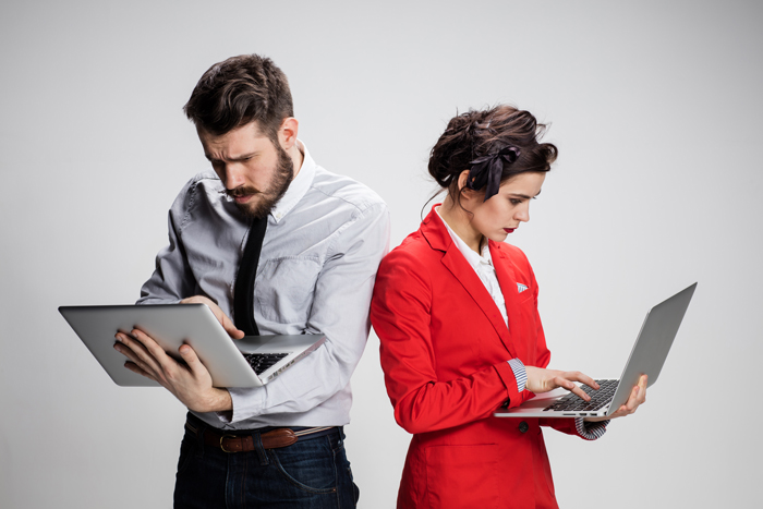 un homme et une femme en tenue d’affaire, debout, tenant chacun un ordinateur portable ouvert à la main, reposant que le bras gauche, semblant chercher avec concentration et angoisse en tapant de la main droite sur le clavier, tête baissée, font plissé.