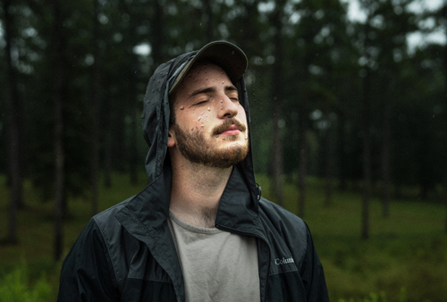 Jeune homme sous la pluie, en veste ouverte imperméable noire à capuche, casquette à visière, les yeux fermés, tête inclinée vers le ciel, le visage barbu offert à la pluie, dans un environnement boisé, clairière entourée d’une forêt.