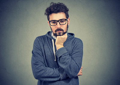 jeune homme à l’air pensif, barbe et lunettes noires, cheveux bouclés mi-courts, sweat décontracté couleur chiné à capuche et ficelle blanche, bras semi-croisés, la main (le poing) sous le menton.