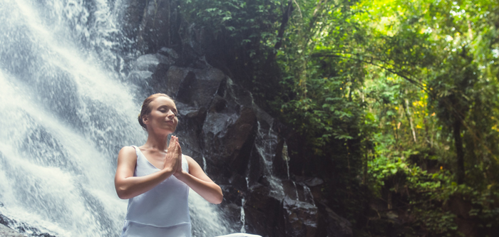 jeune femme paisible, habillée de blanc, cheveux attachés, les yeux fermés, mains jointes en prière ou méditation, sur un rocher, devant une chute d’eau majestueuse, dans un décor naturel sauvage de forêt tropical primaire.