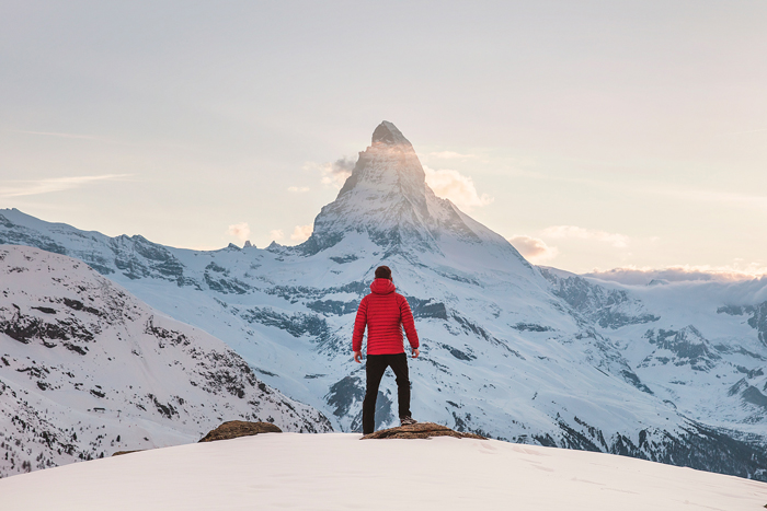 Un homme avec un anorak rouge au sommet des montagnes enneigées observant un pic rocheux