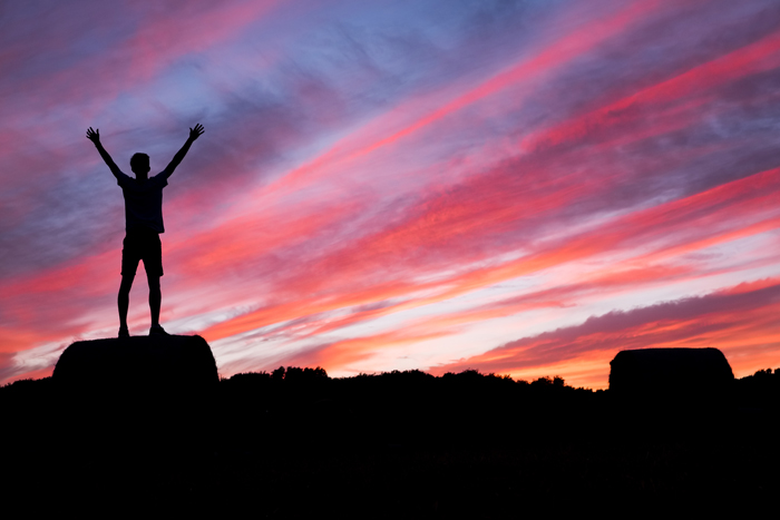 louange; homme levant les mains vers les Cieux, au coucher/lever de soleil aux couleurs chatoyantes, dégradés de bleu, rose, rouge et orange, sous un ciel nuageux; vue de dos, en ombre, dans un milieu naturel.