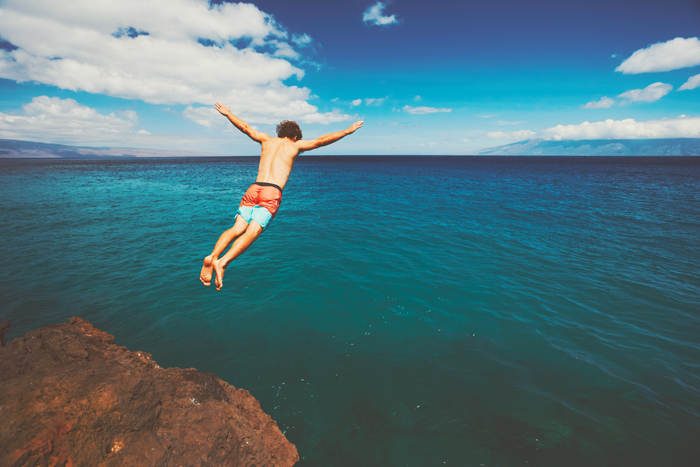 jeune homme les bras ouverts, en caleçon de bain bleu et orange, sautant dans l'océan depuis une falaise