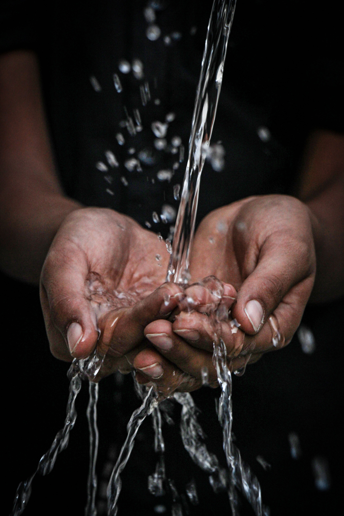 Filet d'eau qui coule dans les mains jointes ouvertes ; gouttes d’eau qui jaillissent et eau qui s’écoule entre les doigts, sur fond noir ; eau vive ; image de la pureté, purification